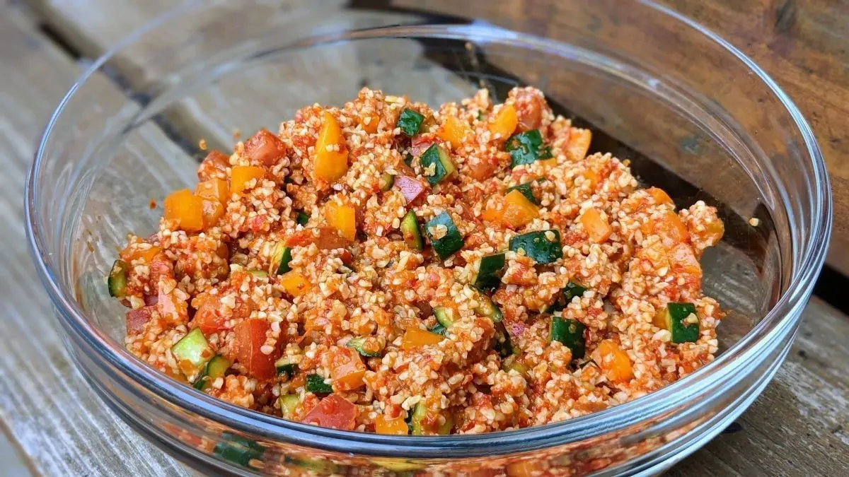 Ein bunter Bulgursalat (Kisir) in einer Glassch&uuml;ssel, frisch zubereitet mit Gem&uuml;se und Ajvar, auf einem Holztisch.