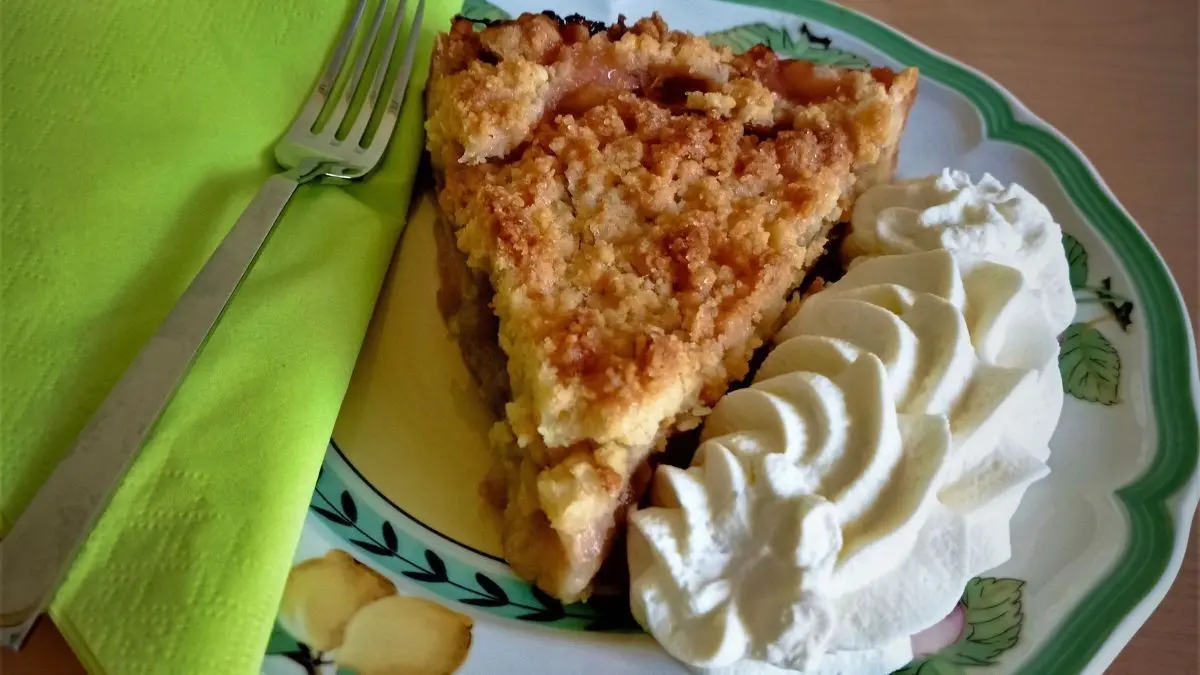 Ein St&uuml;ck Rhabarber-Streusel-Kuchen liegt auf einem Teller, begleitet von Sahne, auf einem Holz-Tisch.