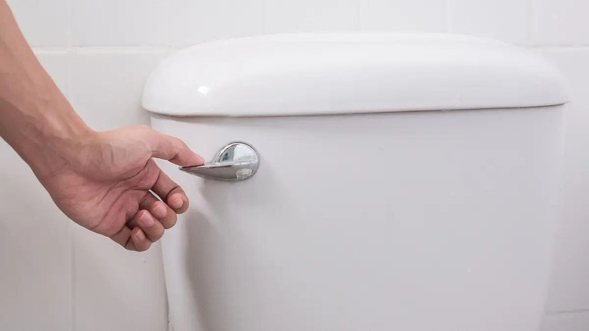 Eine Hand bet&auml;tigt den Sp&uuml;lknopf einer wei&szlig;en Toilette in einem hellen Bad, um Wasser f&uuml;r die Reinigung zu aktivieren.