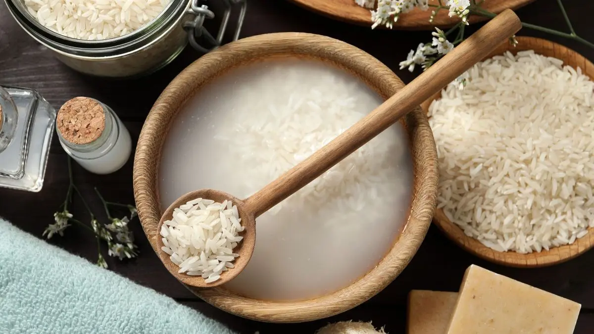 Ein Holzl&ouml;ffel hebt Reiswasser aus einer Schale, umgeben von Reis, einem Handtuch und Naturprodukten f&uuml;r die Haarpflege.