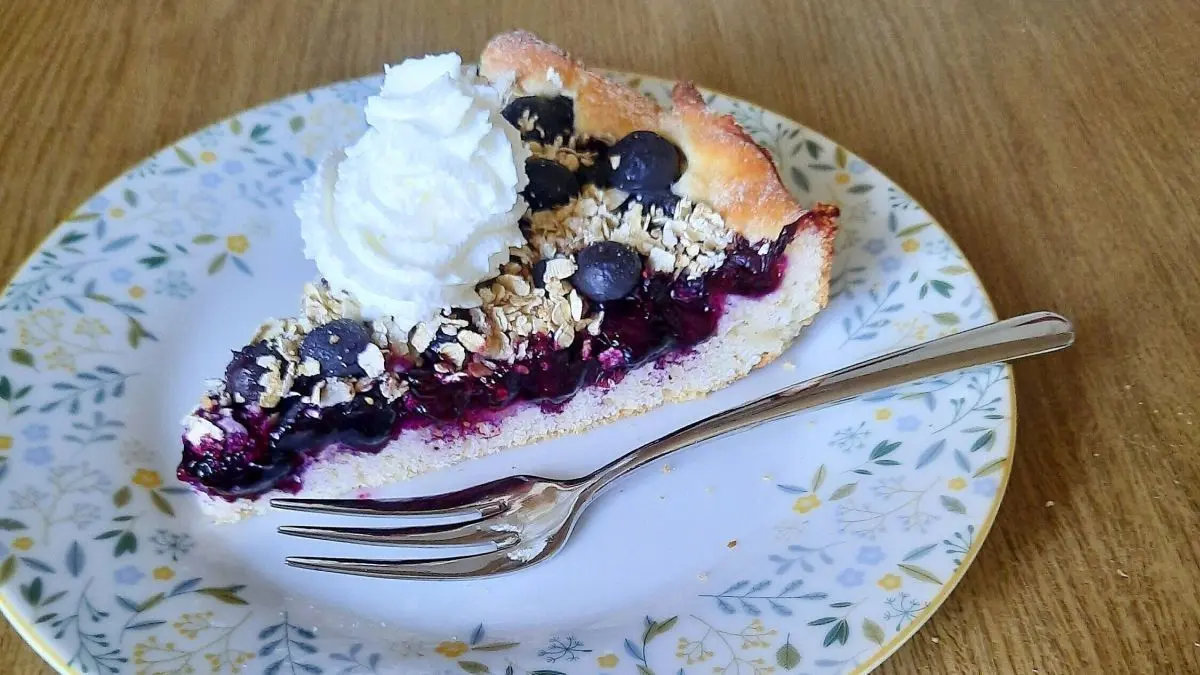 Ein St&uuml;ck Heidelbeerkuchen mit Haferflocken-Crunchy und Sahne auf einem gemusterten Teller steht auf einem Holztisch.