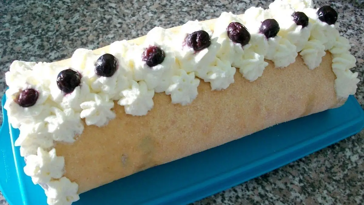 Eine Biskuitrolle liegt auf einem blauen Brett, garniert mit Sahne und Heidelbeeren f&uuml;r einen frischen Genuss.