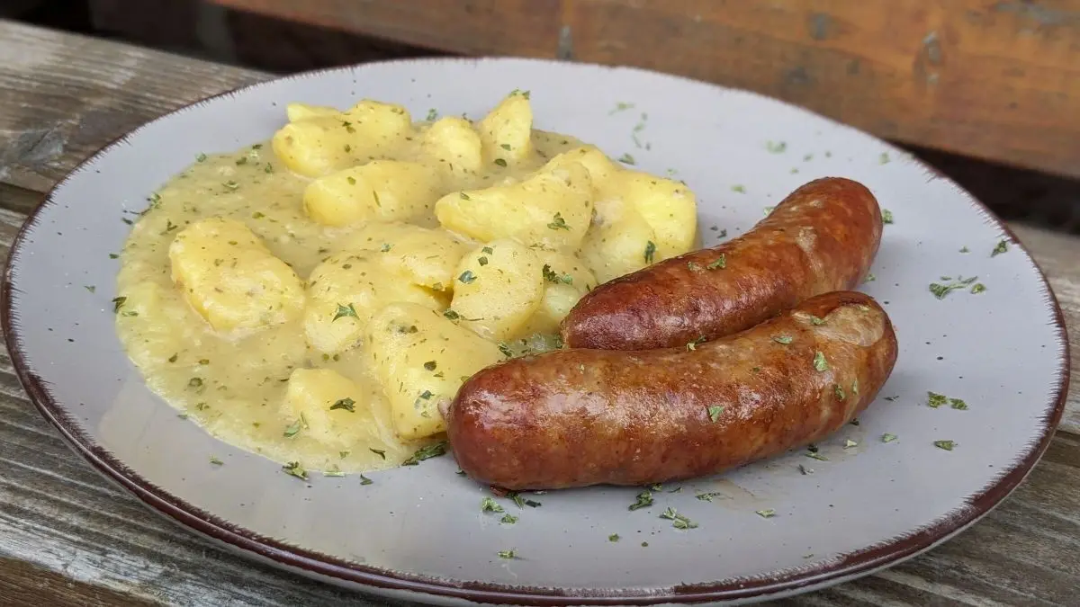Kartoffelgem&uuml;se mit Bratwurst wird appetitlich auf einem wei&szlig;en Teller serviert, umgeben von frischen Kr&auml;utern.