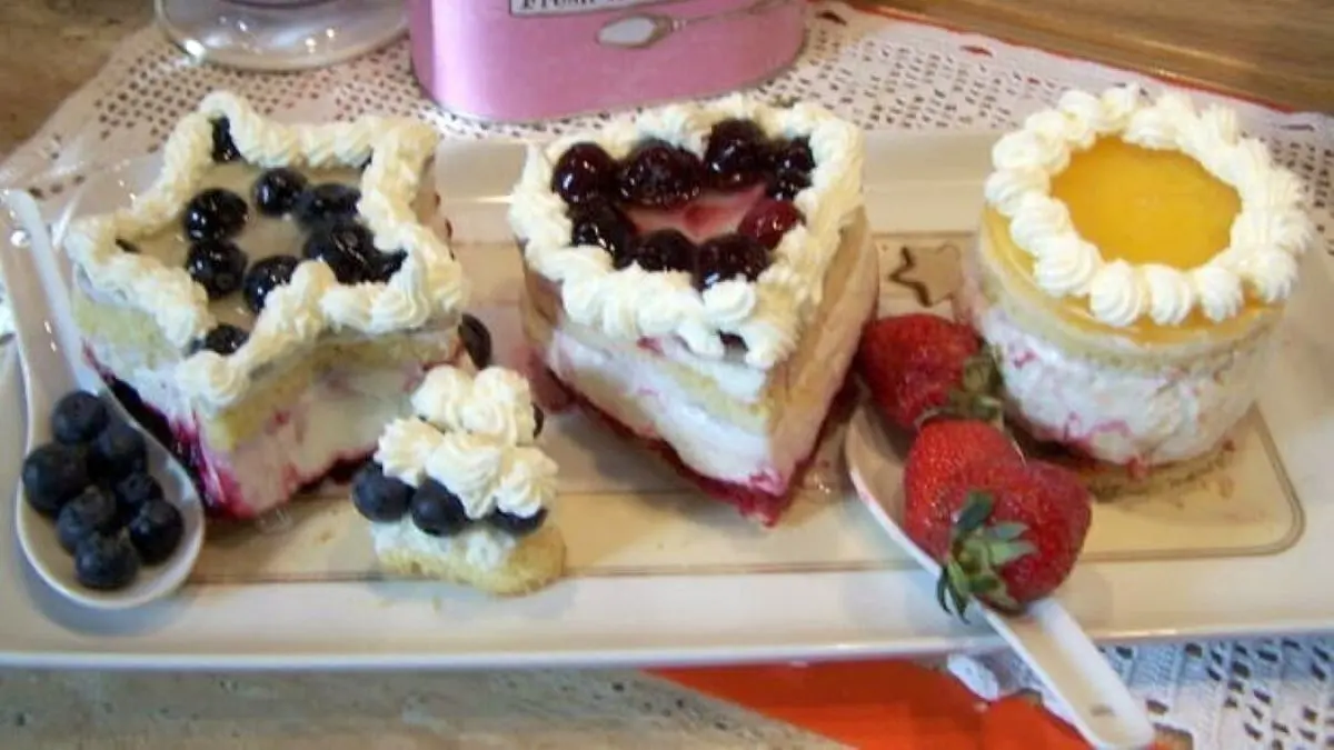 Drei Quark-Sahne-T&ouml;rtchen mit Beeren und Pfirsich stehen auf einem Teller, umgeben von frischen Fr&uuml;chten und einem Kaffeebecher.