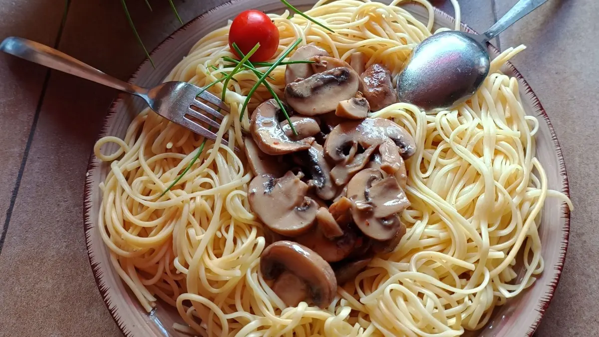 Ein Teller mit Spaghetti wird mit Pilzen garniert, umgeben von roten Tomaten und einem Kr&auml;utertopf.