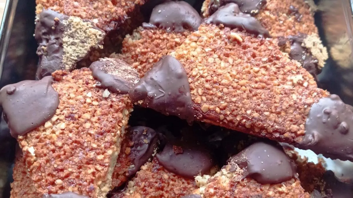Im Fokus sind Nussecken, die in einer metalnen Form angeordnet sind. Sie sind mit einer Schicht Schokolade &uuml;berzogen und liegen in einem hellen Umfeld.
