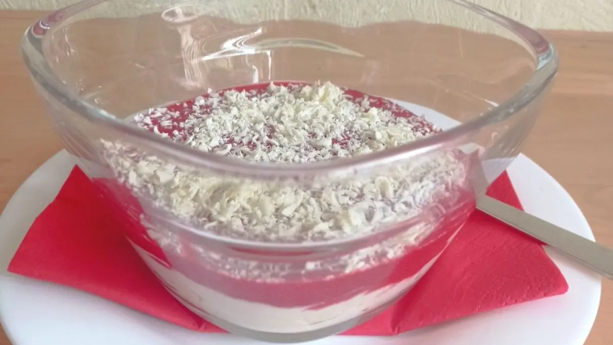 Eine Sch&uuml;ssel mit Himbeer-Dessert liegt auf einem wei&szlig;en Teller, garniert mit Kokosraspeln, auf einem Holztisch.