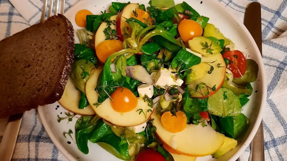 Ein bunter Feldsalat mit Avocado, Apfel und Physalis wird auf einem Tisch serviert, umgeben von einem Brotschnitz.