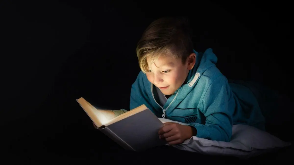 Ein Junge liegt auf einem Kissen und liest gespannt ein Buch, umgeben von Dunkelheit, die die Leselampe betont.