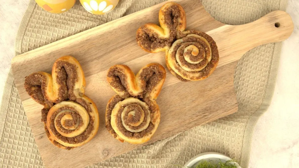 Drei Hefeteig-Osterhasen mit Nussf&uuml;llung liegen auf einem Holzbrett, umgeben von Ostereiern und einem Tuch.