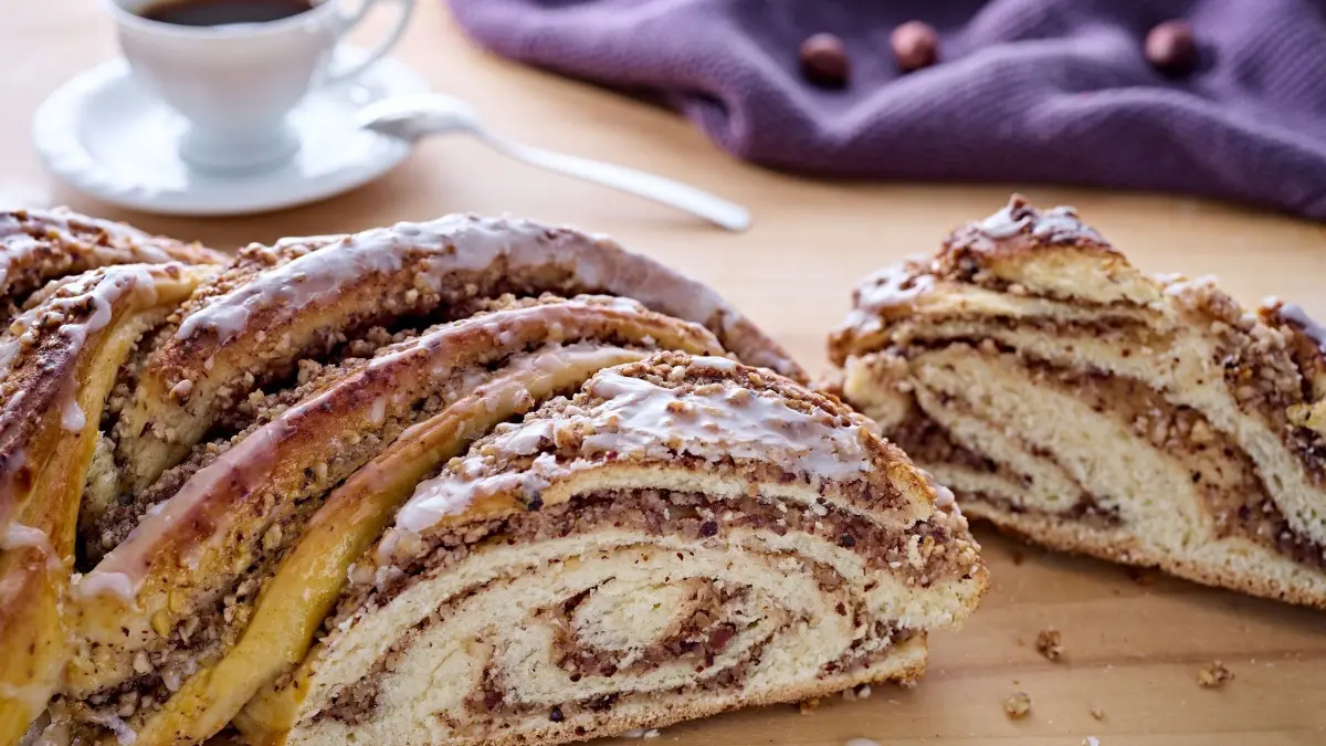 Der Hefezopf mit Nussf&uuml;llung liegt appetitlich auf einem Holztisch, neben einer Tasse Kaffee und einem lila Tuch.
