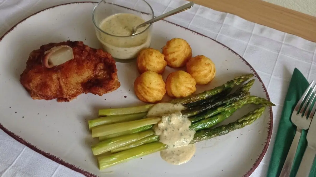 Gr&uuml;ner Spargel und Kalbsschnitzel sind kunstvoll angerichtet, begleitet von Krapfen und einer Sauce auf einem Teller.