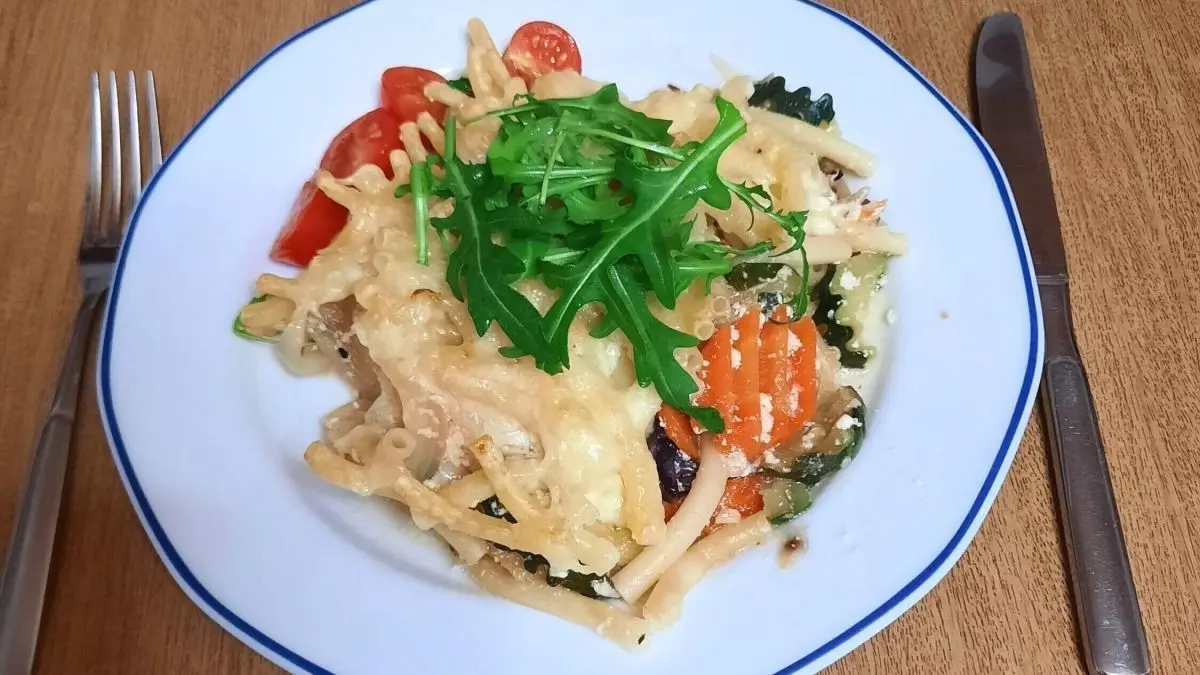 Makkaroni-Gratin liegt auf einem wei&szlig;en Teller, &uuml;bers&auml;t mit buntem Gem&uuml;se und frischem Rucola, auf einem Holztisch.