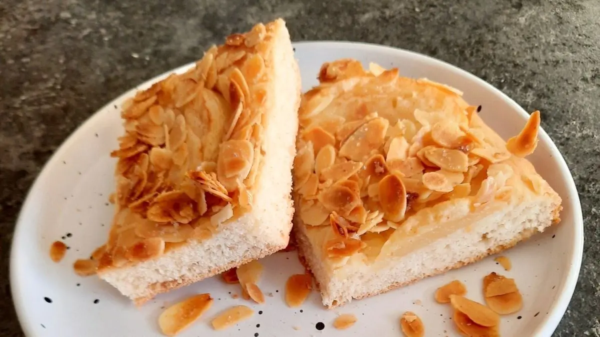 Zwei St&uuml;cke Butterkuchen liegen auf einem wei&szlig;en Teller, bedeckt mit ger&ouml;steten Mandeln, umgeben von grauem Tisch.