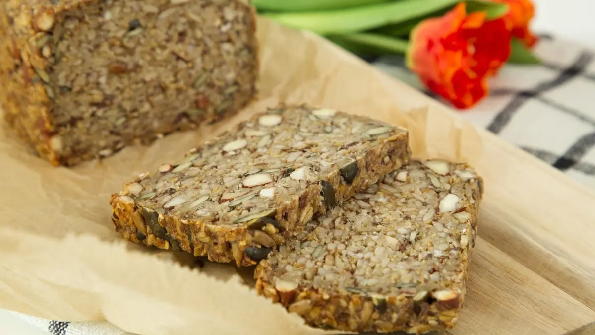 Ein Brot aus Saaten liegt auf einem Holzbrett, w&auml;hrend frische Tulpen im Hintergrund sichtbar sind.