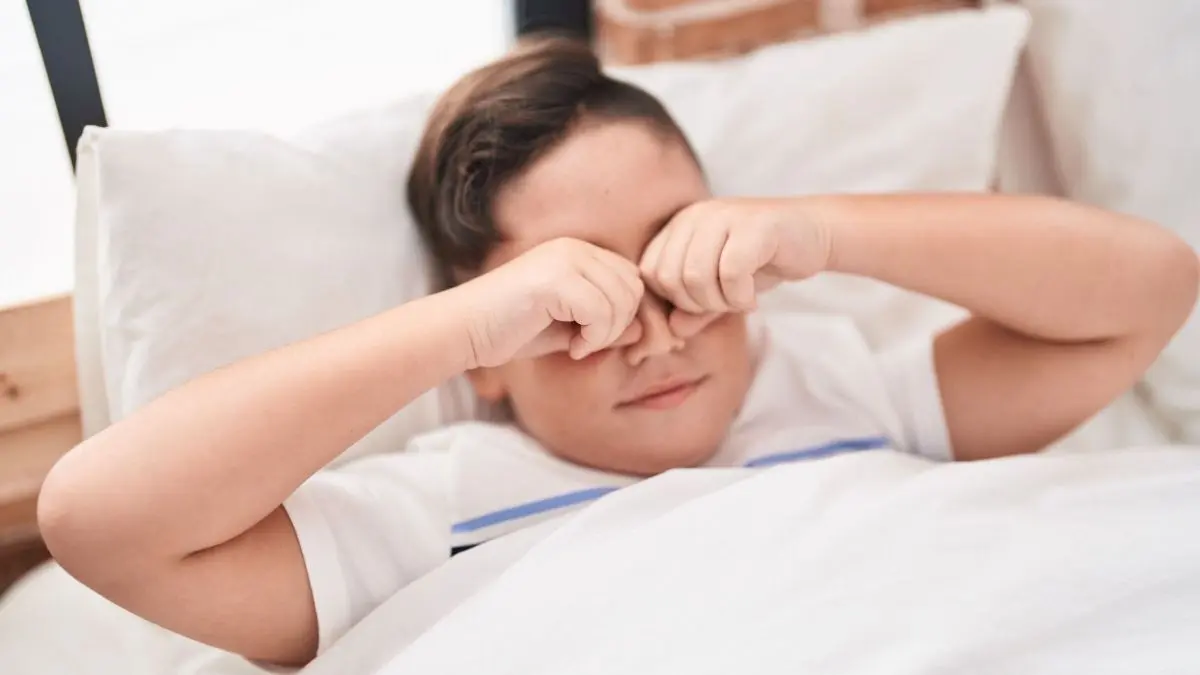 Ein Junge reibt sich die Augen, w&auml;hrend er im Bett liegt und frisch aufwacht, um den Schlafsand zu vertreiben.
