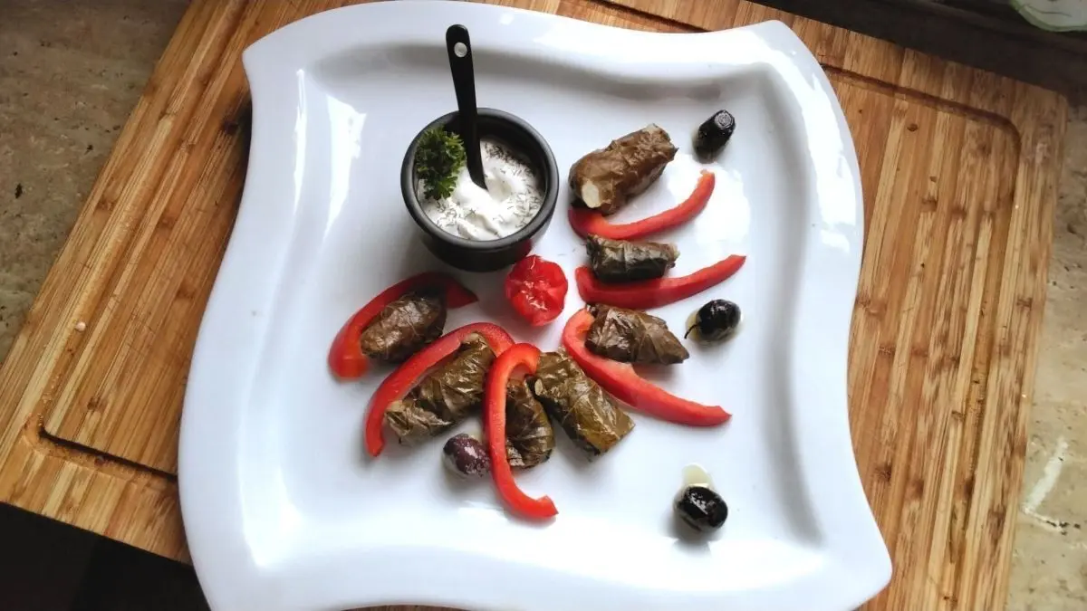 Gef&uuml;llte Weinbl&auml;tter liegen auf einem wei&szlig;en Teller, umgeben von Joghurt Dip, Paprika und Oliven auf einem Holztisch.
