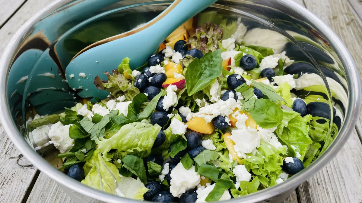 Der Salat mit Blaubeeren, Feta, Pfirsichen und Basilikum liegt in einer silbernen Sch&uuml;ssel auf einem Holztisch.