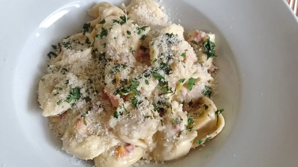 Tortelloni liegen in einer cremigen Speck-Sahne-So&szlig;e, bestreut mit K&auml;se und Kr&auml;utern, in einem wei&szlig;en Teller auf Holztisch.