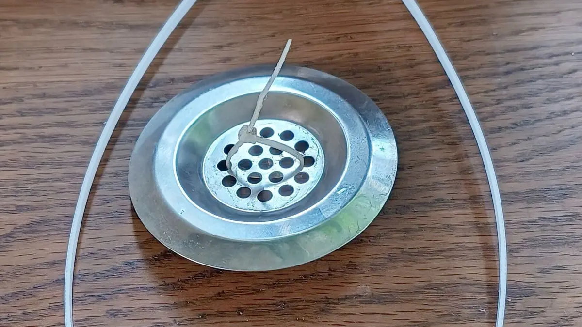 Ein metallisches Abflusssieb liegt auf einem Holztisch, fixiert mit einem Kabelbinder. Ein Zahnstocher steckt in den L&ouml;chern. Text: 17. Sept. 2024, 13:28.