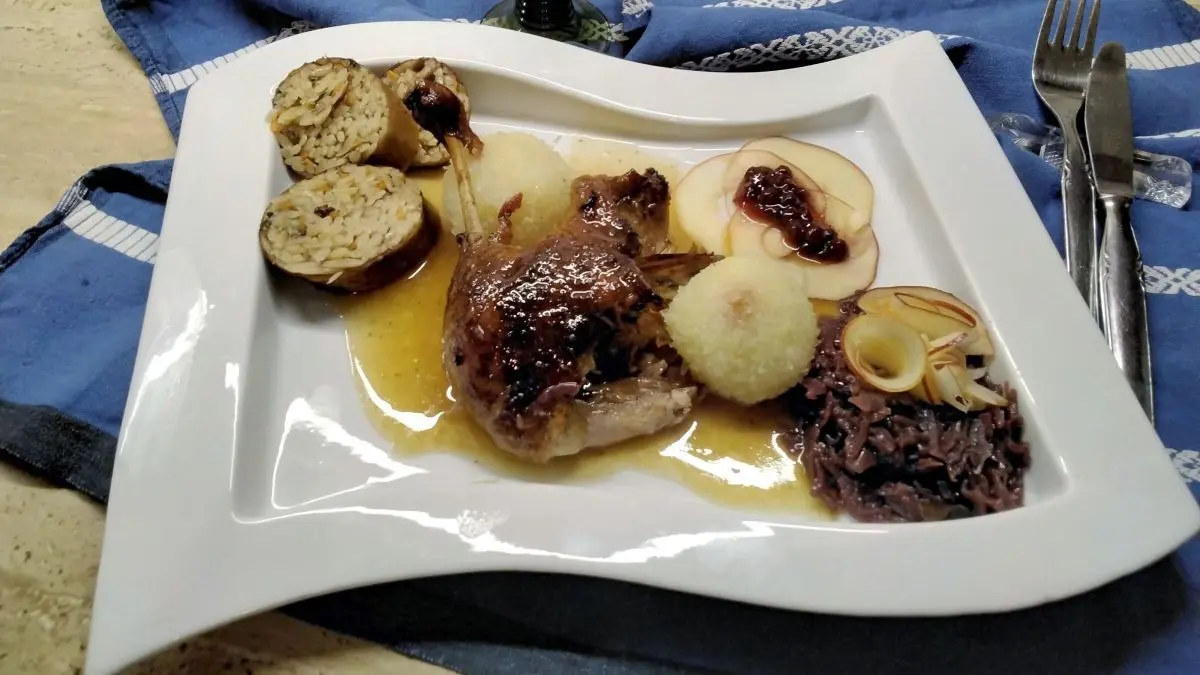 Gericht auf wei&szlig;em Teller: Entenhals gef&uuml;llt mit Kritharaki, dazu So&szlig;e, Kn&ouml;del, Apfelscheiben, Rotkohl; im Hintergrund Rotwein.