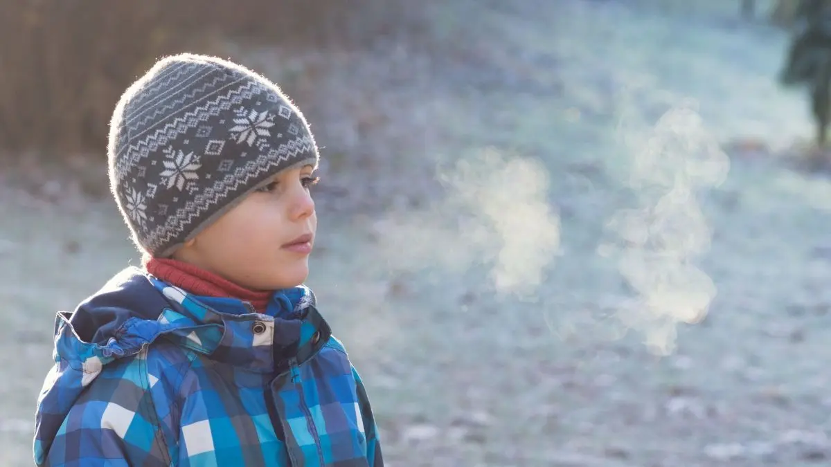 Ein Kind mit M&uuml;tze atmet im Freien; der Atem ist sichtbar in der kalten Winterluft auf einem frostigen Rasen.