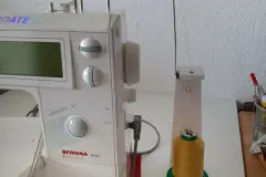 Garnrollenhalter samt Garn wird neben die Maschine gestellt und der Metall-Arm so abgewinkelt, dass das Garn in gleicher H&ouml;he wie (im weiteren Verlauf) der entsprechende Einzug in die N&auml;hmaschine vorgegeben ist.