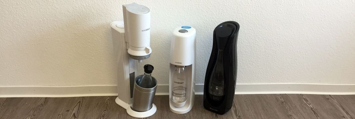 Ein Wassersprudler steht auf einem Holzboden, bereit, Wasser zu sprudeln. Daneben stehen zwei Flaschen für karbonisiertes Wasser.