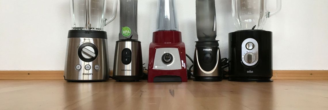 In einer Reihe stehen fünf Smoothie-Maker auf einem Holzboden, bereit, frische Getränke zuzubereiten.