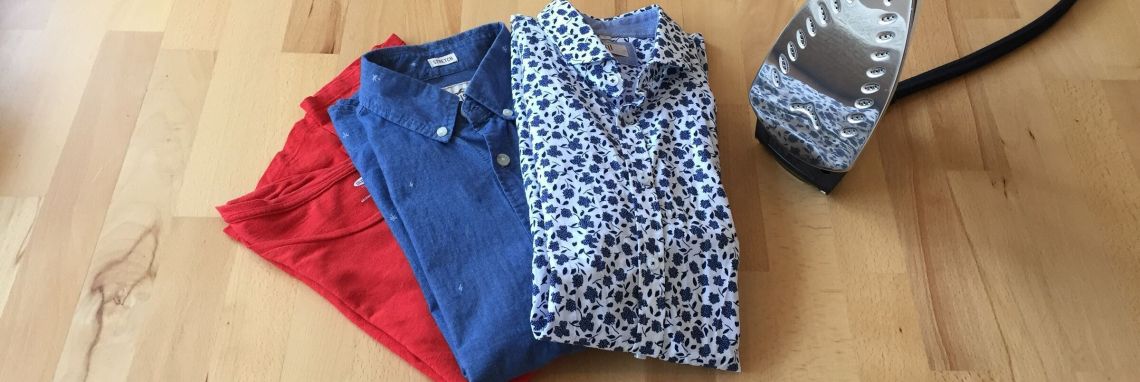 Ein Dampfbügeleisen steht neben einem Stapel gefalteter Kleidung in Rot, Blau und mit Muster auf einem Holzboden.